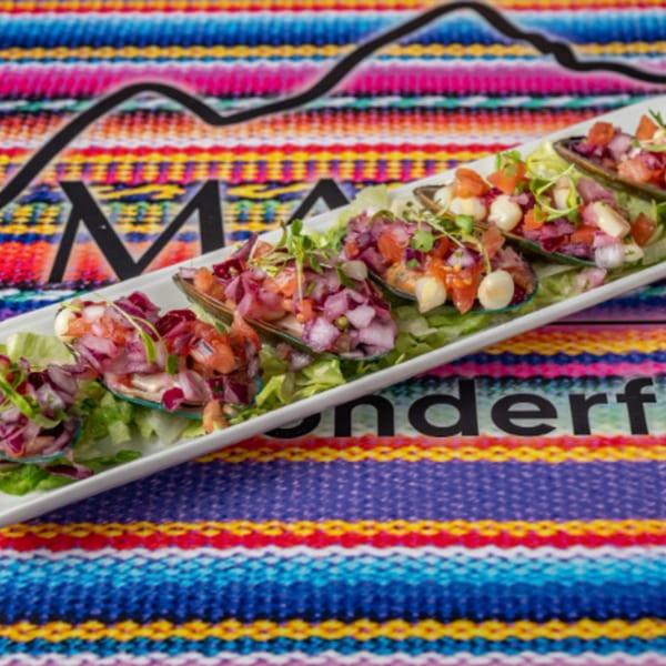 Peruvian mussels topped with red onion, corn, and tomato on a white platter over a colorful striped tablecloth.