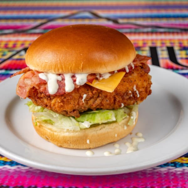 Crispy chicken sandwich with bacon, cheese, lettuce, and white sauce on a bun, on a white plate.