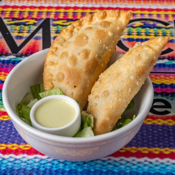 Two golden-brown Peruvian empanadas with a side of green sauce and lettuce in a white bowl.