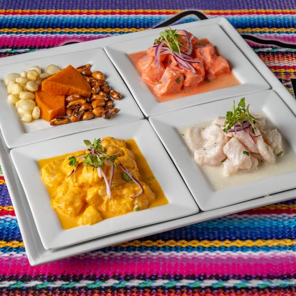 Four square plates of Peruvian ceviche and sides, including corn and sweet potato, on a colorful striped mat.